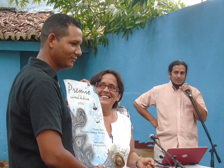 A la izquierda, Alexander Jiménez del Toro, al recibir el Premio Toda luz y toda mía por su obra Colmillos de costa, de manos de Merari Mangly. Al fondo, Karel Leyva, integrante del jurado. Foto: Tomada del periódico Escambray